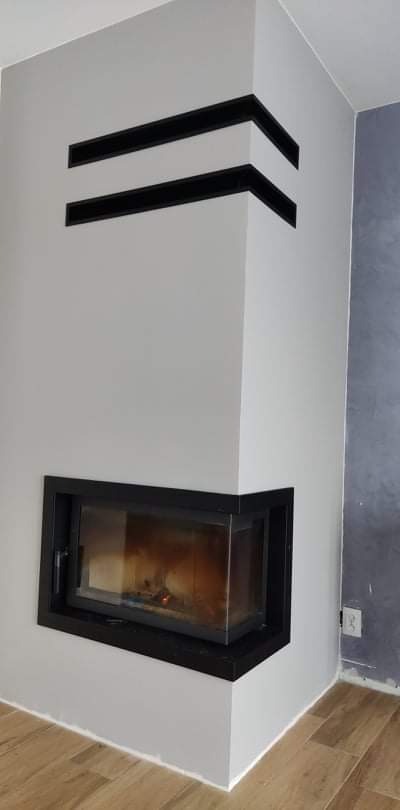 Nowoczesny kominek narożny z czarną obudową i dwoma poziomymi, czarnymi wnękami dekoracyjnymi w szarej ścianie, widoczny fragment drewnianej podłogi.