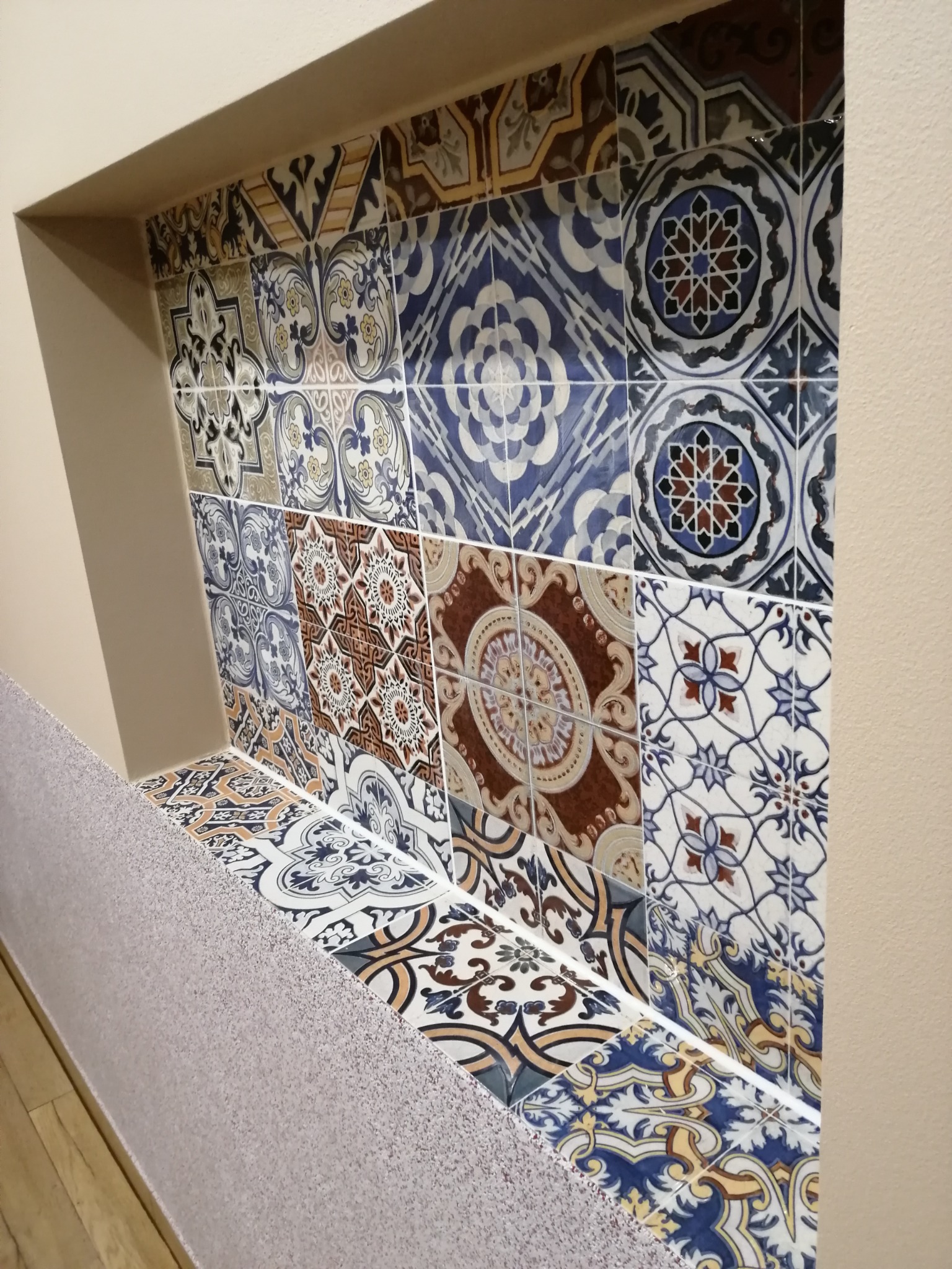 Nisza ścienna wyłożona dekoracyjnymi, wzorzystymi płytkami ceramicznymi w odcieniach błękitu, brązu i beżu, z widocznym fragmentem drewnianej podłogi i szarego parapetu.
