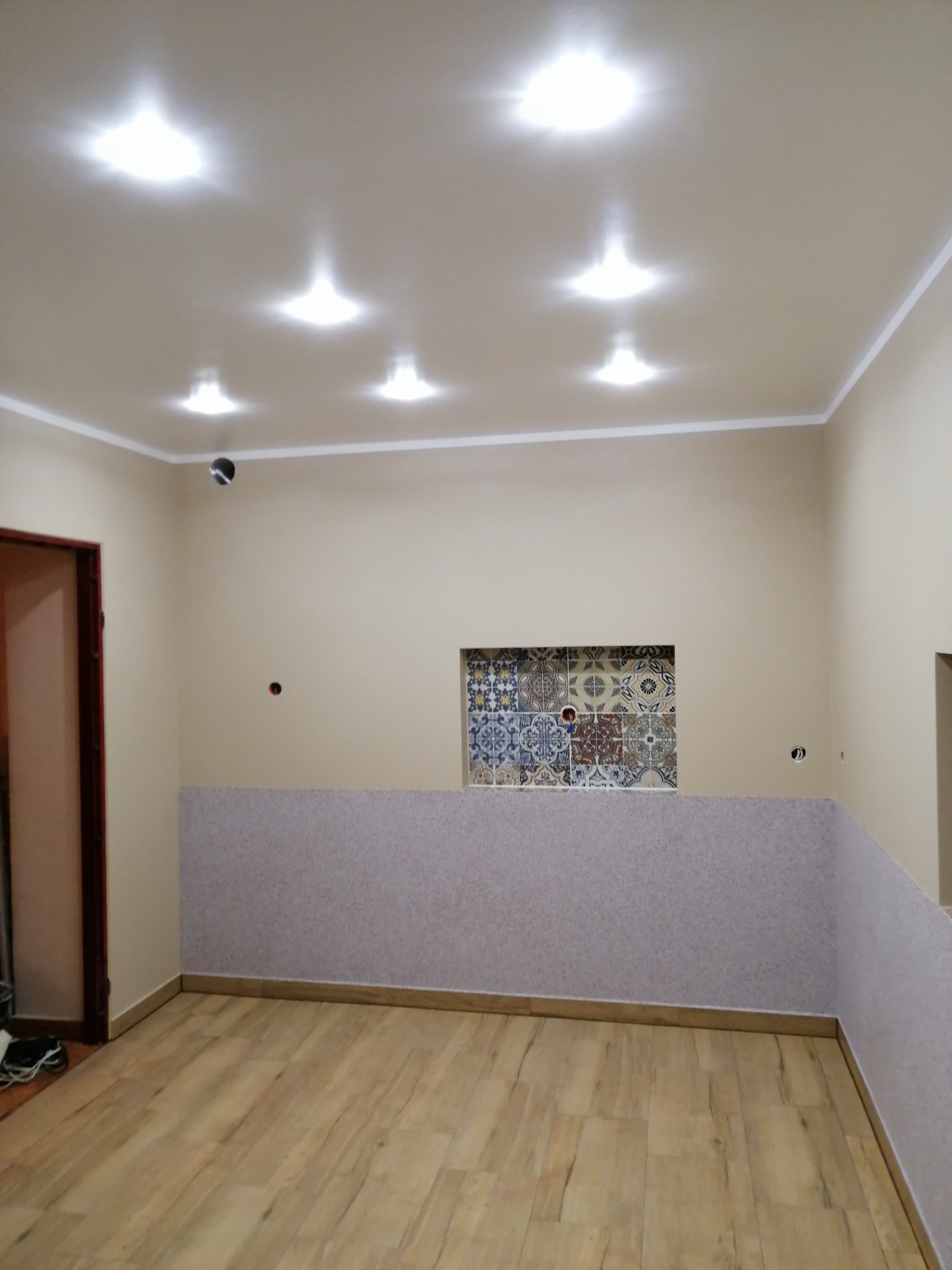 Wnętrze pokoju po częściowym remoncie: położone panele podłogowe, fragment ściany pokryty tapetą, dekoracyjna wnęka z mozaiką, widoczne punkty świetlne w suficie podwieszanym.