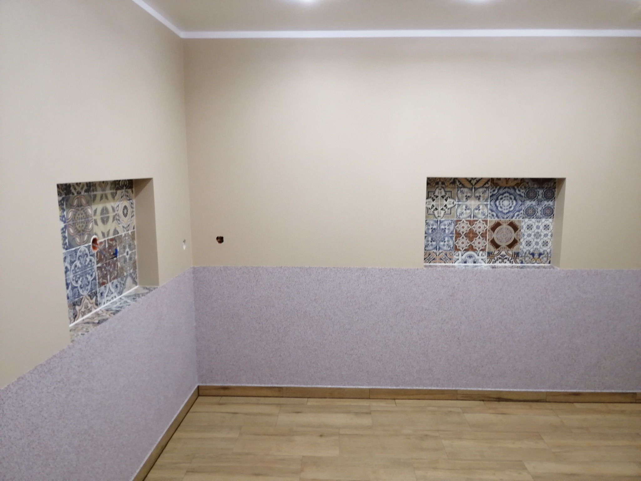 Wnęki z dekoracyjnymi, wzorzystymi płytkami ceramicznymi w ścianie pomalowanej na beżowo, dolna część ściany pokryta tapetą strukturalną, jasna podłoga z paneli imitujących drewno.