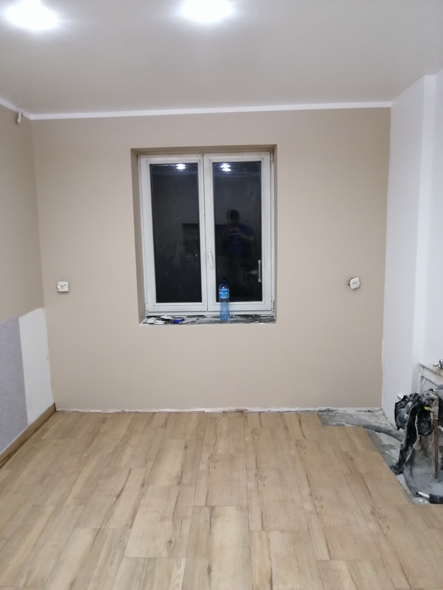 Pokój po częściowym ułożeniu paneli podłogowych, ściany pomalowane na beżowo, widoczne gniazdka elektryczne i butelka wody na parapecie.