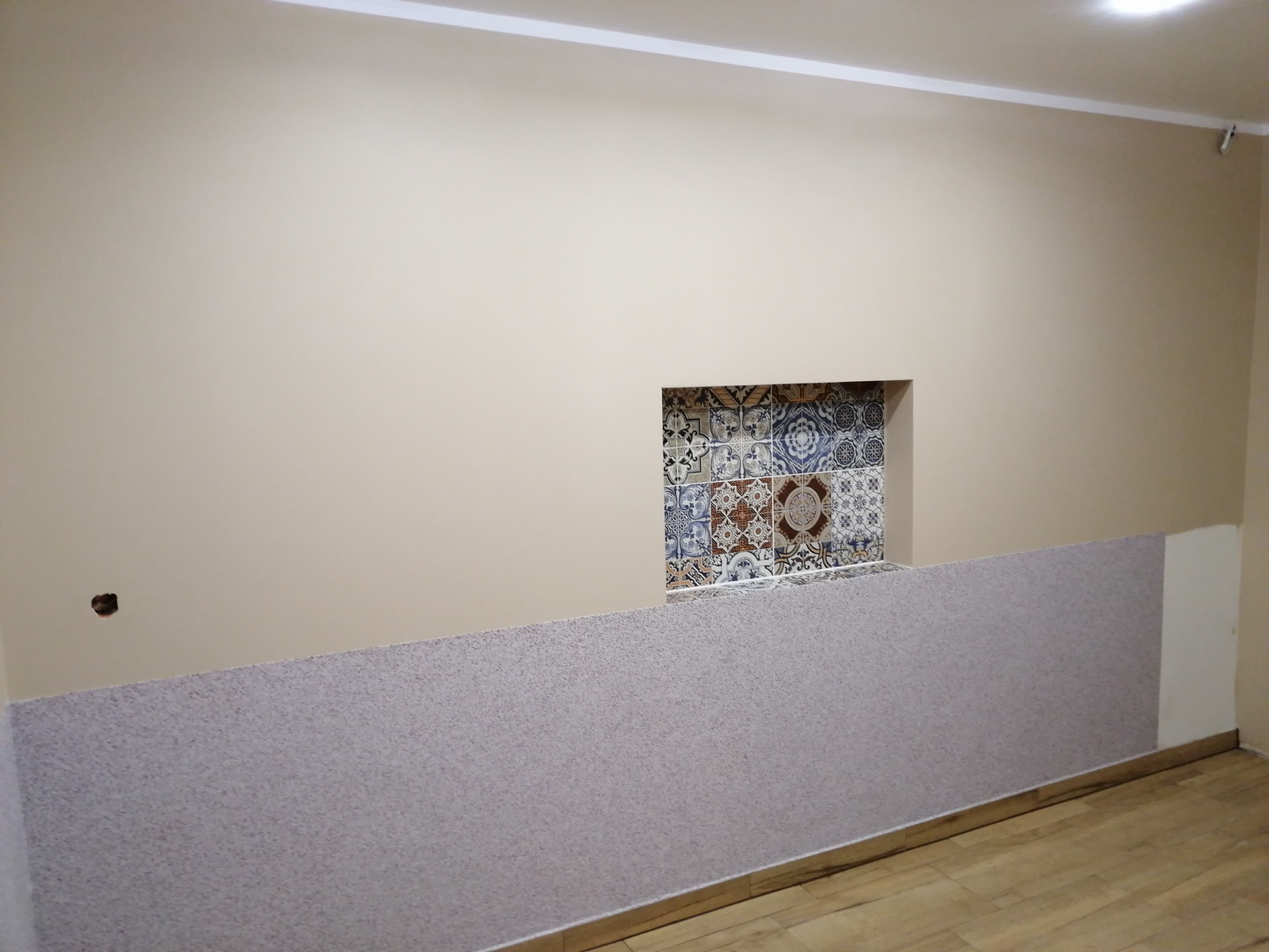 Fragment ściany po remoncie z dekoracyjną wnęką wyłożoną mozaiką, dolna część pokryta szarą tapetą strukturalną, widoczny fragment parkietu.