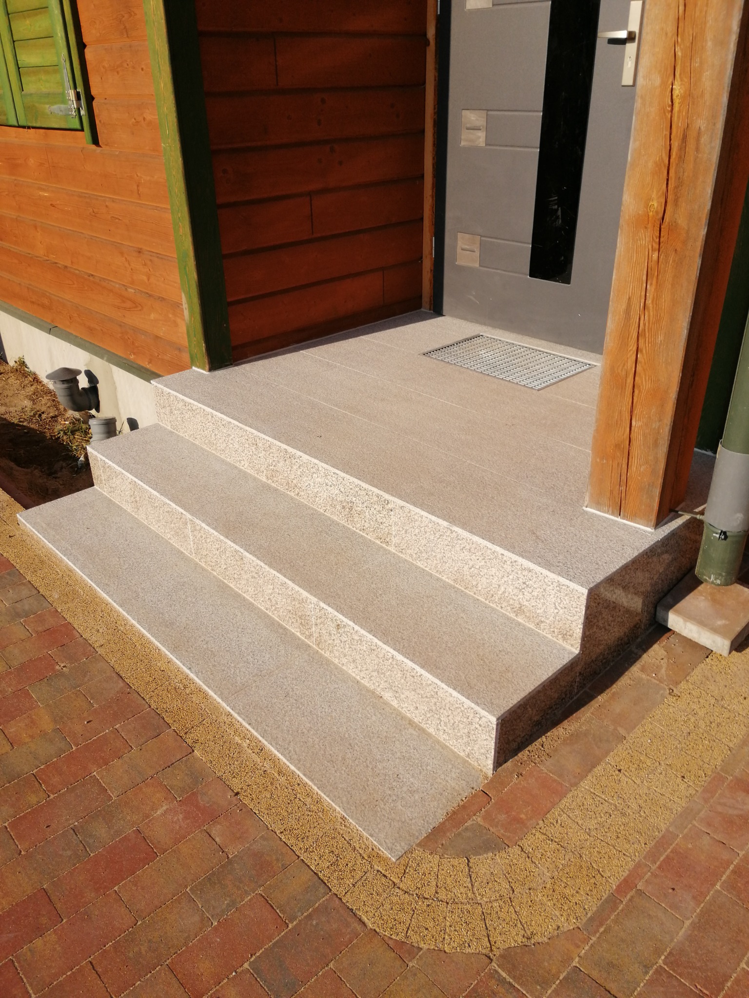 Granitowe schody wejściowe z trzema stopniami, obramowane żywicznym dywanem i czerwoną kostką, prowadzące do szarych drzwi z drewnianym filarem i kratką ściekową.