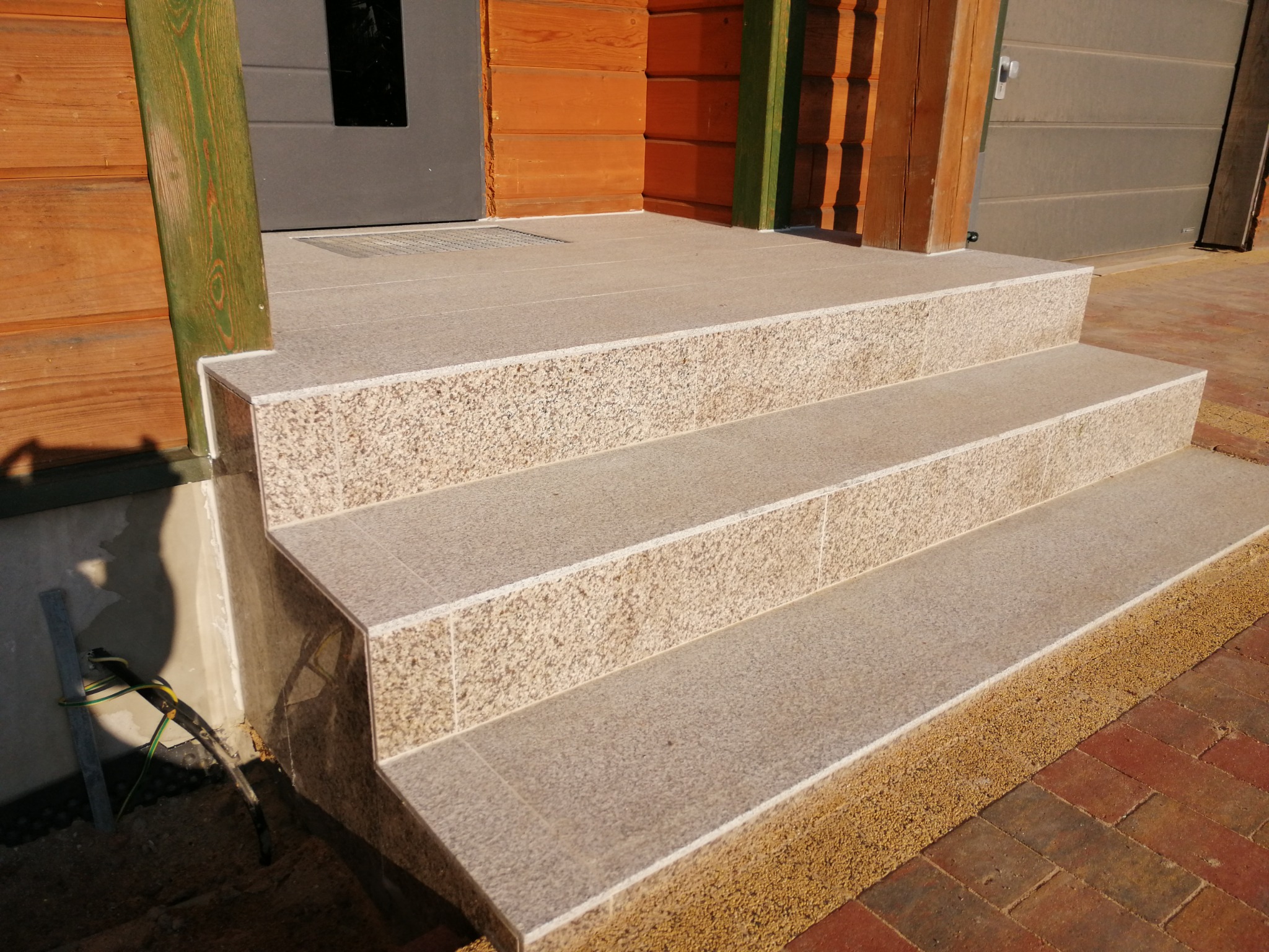 Schody wejściowe wyłożone jasnymi płytkami granitowymi, widoczne przewody elektryczne wystające spod schodów.