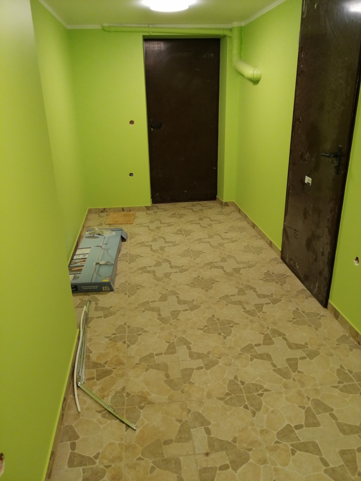 Korytarz z zielonymi ścianami i brązowymi drzwiami, na podłodze płytki imitujące kamień. W rogu leży karton z zestawem prysznicowym i listwy przypodłogowe.