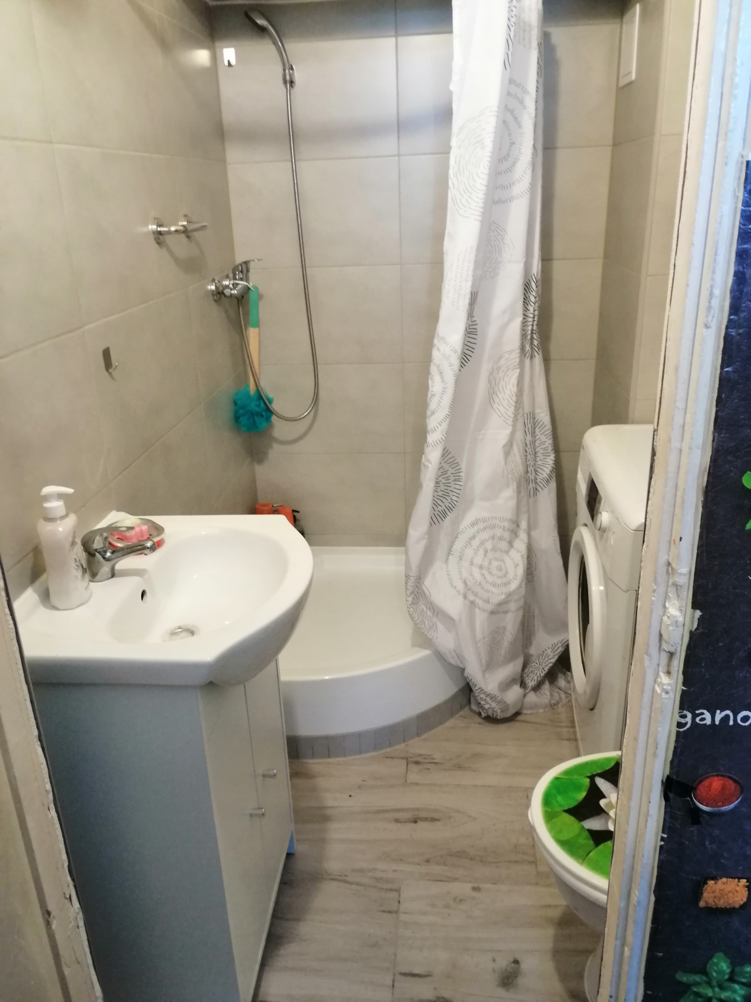 Niewielka łazienka w Suwałkach z kabiną prysznicową, umywalką z szafką, pralką i zasłoną prysznicową w biało-szare wzory. Na ścianie widoczny zestaw prysznicowy i akcesoria łazienkowe.