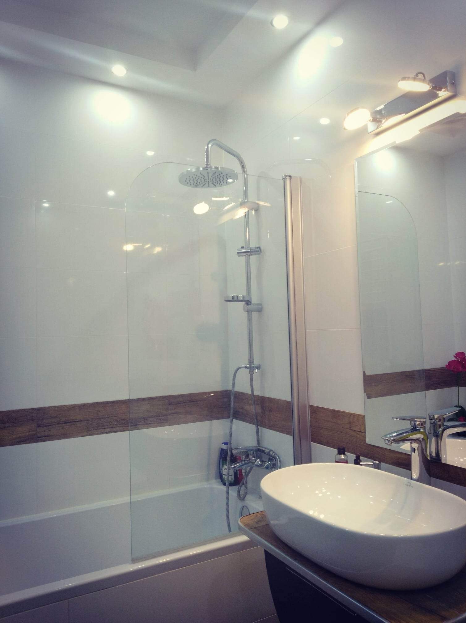 Nowoczesna łazienka z białą armaturą, kabiną prysznicową ze szklanym panelem i drewnopodobnym akcentem na ścianach.