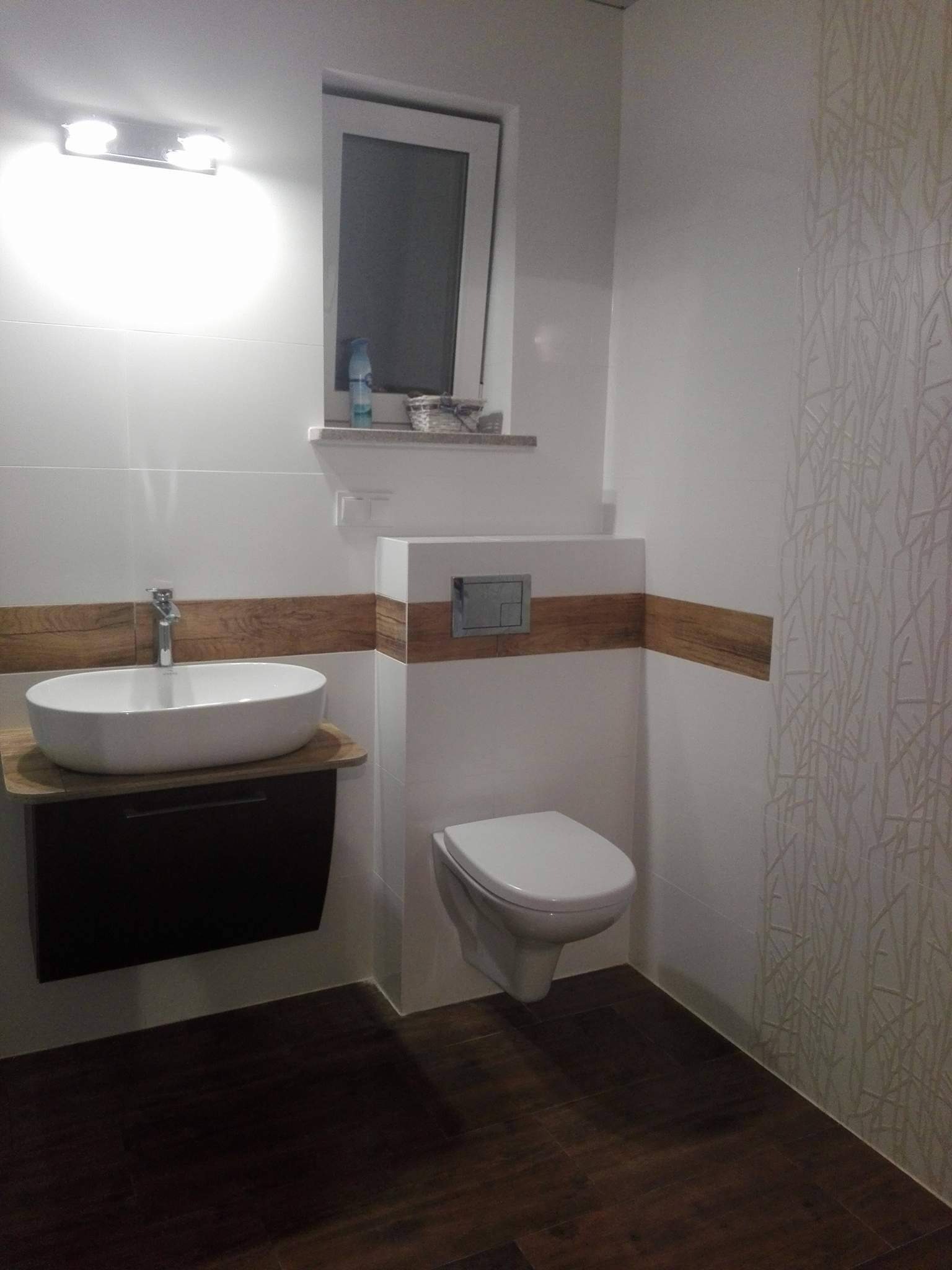 Nowoczesna łazienka z owalną umywalką nablatową na ciemnej szafce, wiszącą toaletą z chromowanym przyciskiem spłukiwania, drewnopodobnymi akcentami i płytkami z subtelnym wzorem roślinnym.