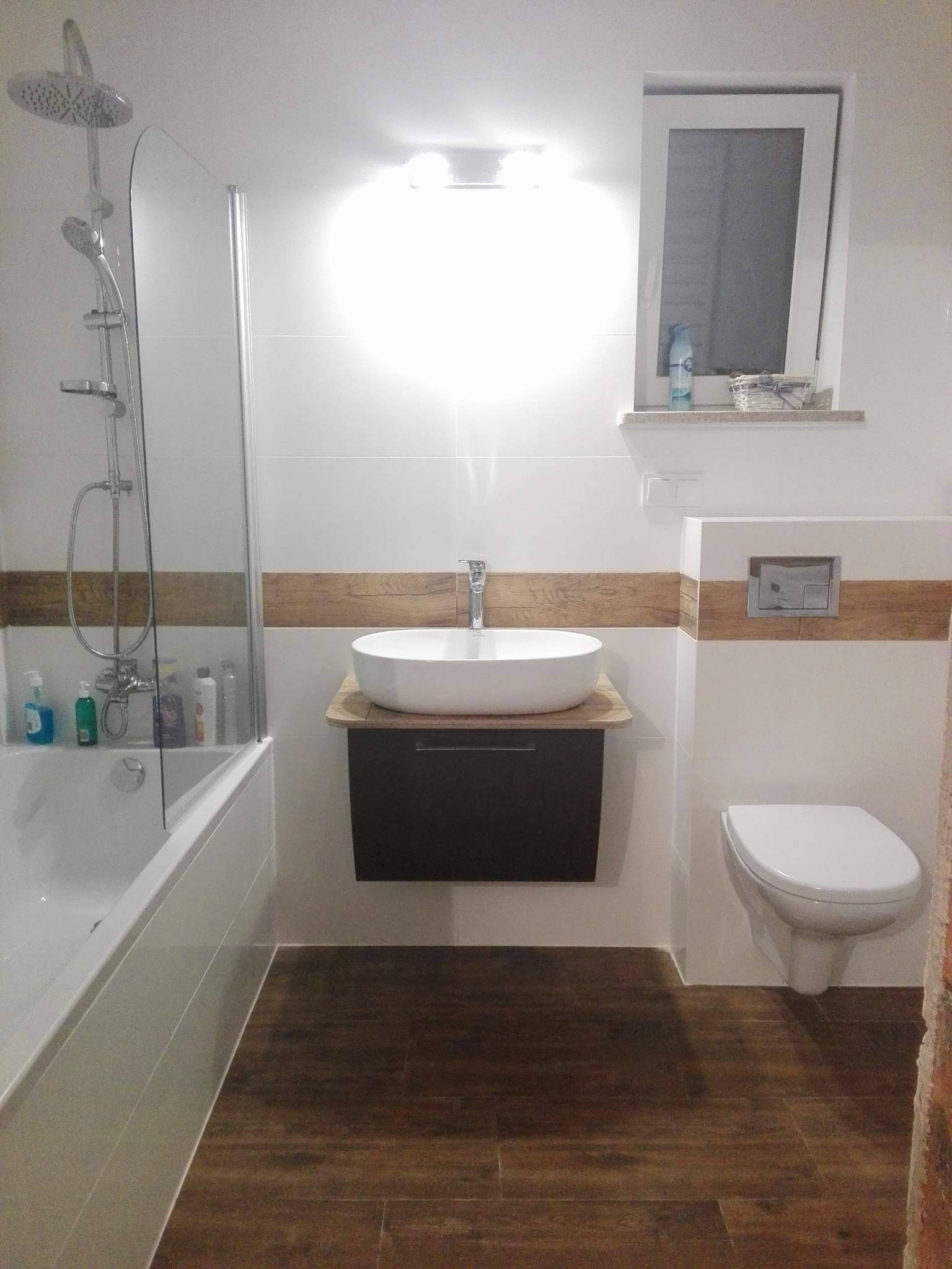 Nowoczesna łazienka z białymi płytkami, drewnopodobnymi akcentami, owalną umywalką nablatową na szafce w kolorze ciemnego brązu, wiszącą toaletą i kabiną prysznicową ze szklanym panelem.