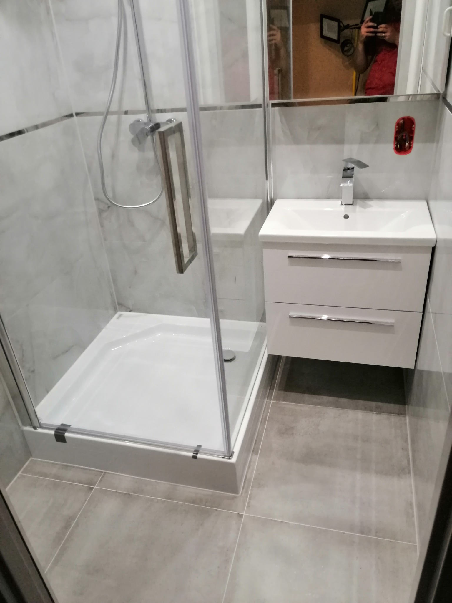 Nowoczesna łazienka z kabiną prysznicową, białą umywalką z szafką i szarymi płytkami podłogowymi, widoczne odbicie osoby robiącej zdjęcie w lustrze.