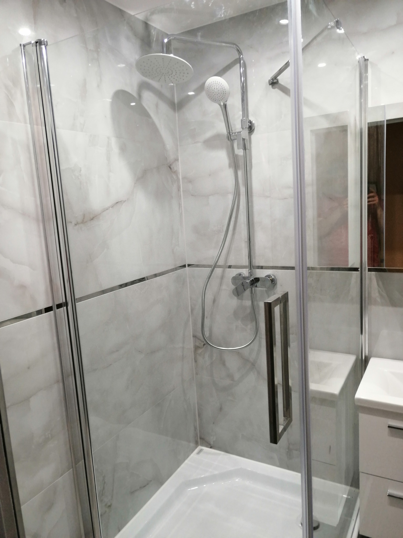 Nowoczesna kabina prysznicowa z deszczownicą i słuchawką, wykończona płytkami imitującymi marmur z metalowymi listwami dekoracyjnymi, widoczna umywalka z szafką w tle.