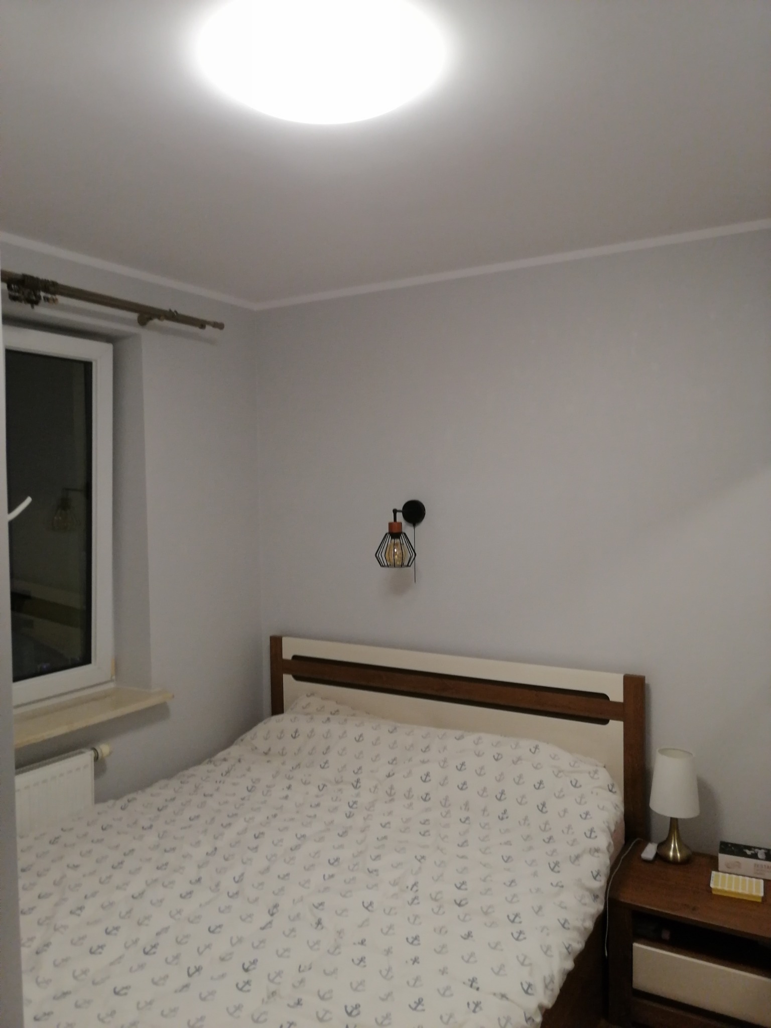 Jasna sypialnia z łóżkiem w pościeli z motywem kotwic, lampką nocną na drewnianym stoliku i nowoczesnym kinkietem na szarej ścianie.