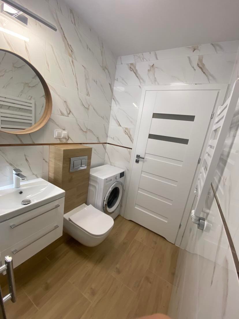Nowoczesna łazienka z marmurowymi płytkami ściennymi, drewnopodobną podłogą, okrągłym lustrem i podwieszaną toaletą obok pralki.