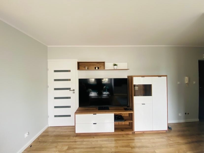 Nowoczesny salon z drewnianą podłogą, szarą ścianą i białymi meblami: szafka RTV z białymi szufladami, szafka z witryną i półki wiszące z dekoracjami.