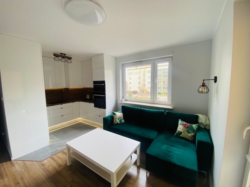 Nowoczesne wnętrze z aneksem kuchennym, białymi meblami na wysoki połysk, drewnianym blatem i oświetleniem LED, szarą podłogą, zieloną sofą narożną i białym stolikiem kawowym przy oknie.