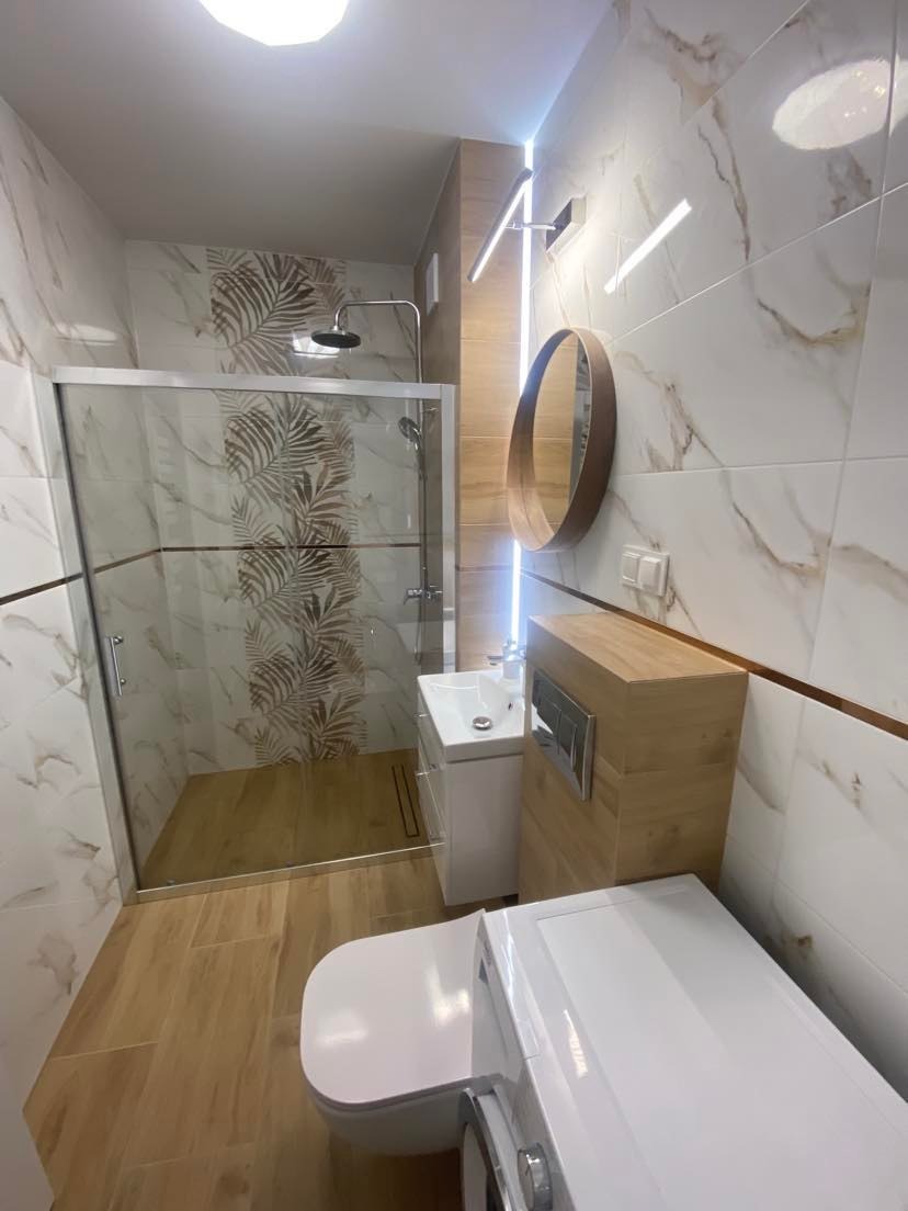 Nowoczesna łazienka z kabiną prysznicową typu walk-in, płytkami imitującymi marmur z motywem liści, okrągłym lustrem i drewnianą szafką pod umywalką, oświetleniem LED oraz pralką w rogu.