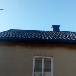 Dom z ciemną dachówką karpiówką, czarną rynną i kominem na tle błękitnego nieba; widoczna antena telewizyjna i numer posesji.