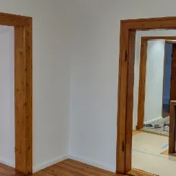 Białe ściany i drewniane futryny drzwiowe w odnowionym wnętrzu. Widoczna podłoga z desek i gniazdko elektryczne na ścianie. W głębi widoczne dalsze pomieszczenia i schody z drewnianą balustradą...