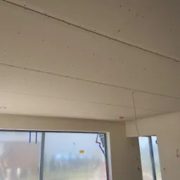 Montaż płyt gipsowo-kartonowych na suficie w nowym budynku, widoczne łączenia płyt i ślady po wkrętach, okno zabezpieczone folią malarską.