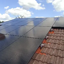 Częściowy widok paneli słonecznych na dachu z czerwonej dachówki, na tle błękitne niebo z białymi obłokami, widoczna antena satelitarna.