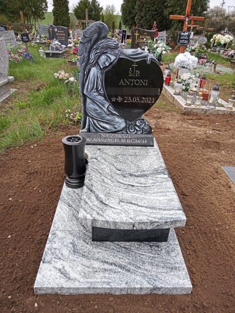 Czarny nagrobek z rzeźbą anioła klęczącego przy sercu z wygrawerowanym imieniem i datą, na tle cmentarza.