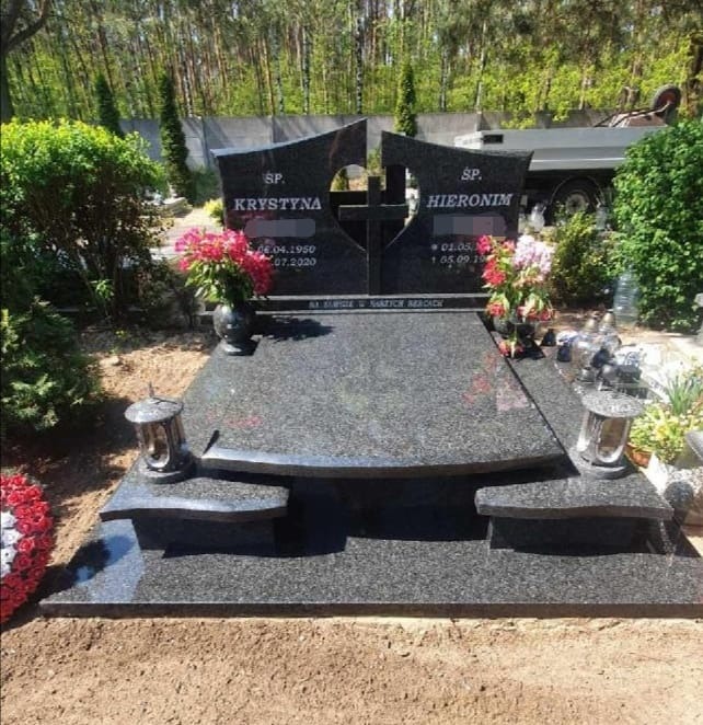 Podwójny, ciemny nagrobek z granitu z wyciętym krzyżem pośrodku, ozdobiony kwiatami w wazonach i lampionami, na tle zieleni cmentarza i fragmentu ciężarówki.