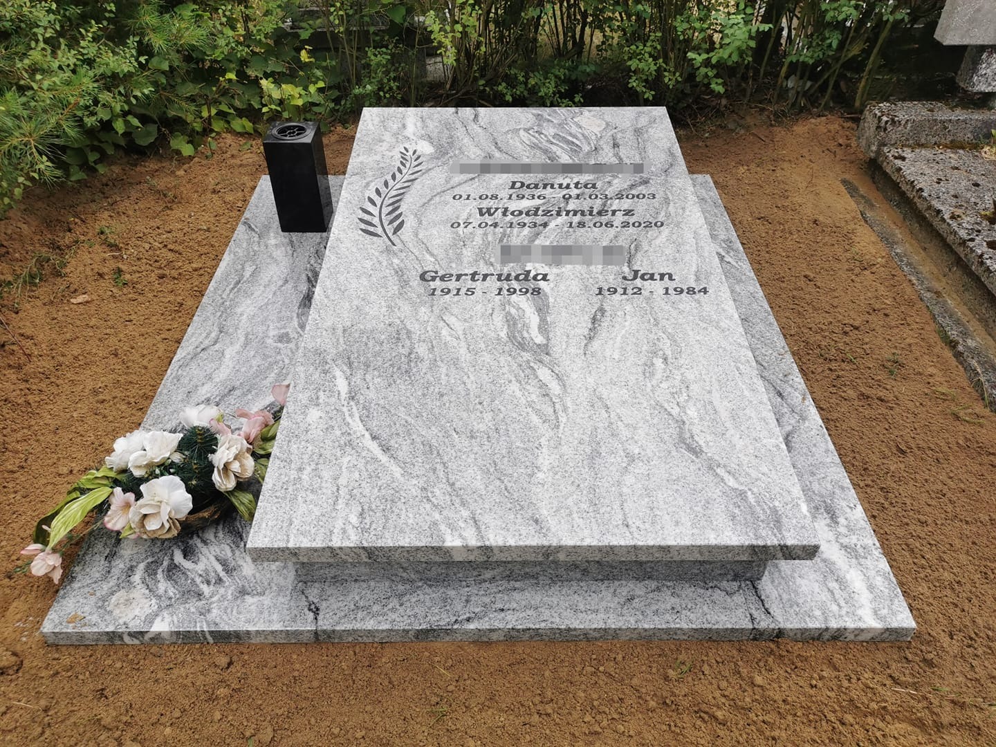 Dwupoziomowy, jasnoszary pomnik z granitu z wygrawerowanymi imionami i datami, czarny wazon na kwiaty i wiązanka białych kwiatów u podstawy, świeżo usypana ziemia wokół.