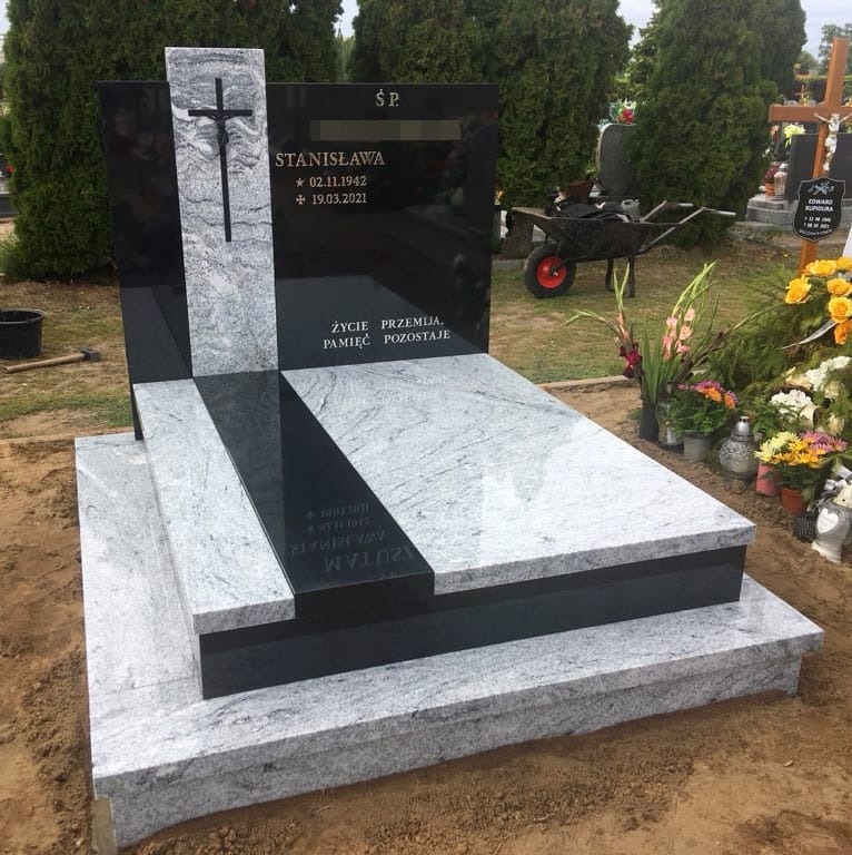 Granitowy pomnik z krzyżem, czarno-biały, z wygrawerowanymi danymi osoby zmarłej, otoczony zielenią cmentarza i kwiatami.
