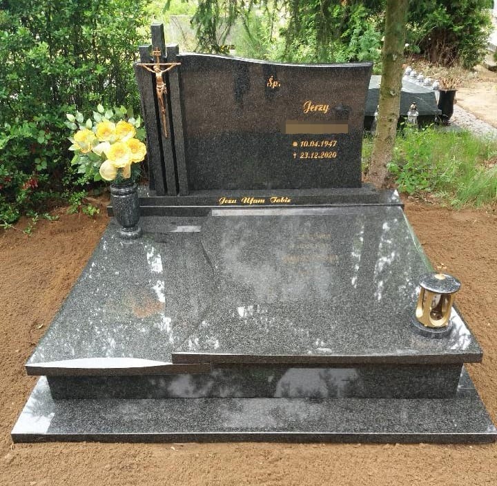 Granitowy pomnik z krzyżem i figurą Jezusa, wazonem z żółtymi kwiatami oraz lampionem, otoczony świeżo usypaną ziemią.