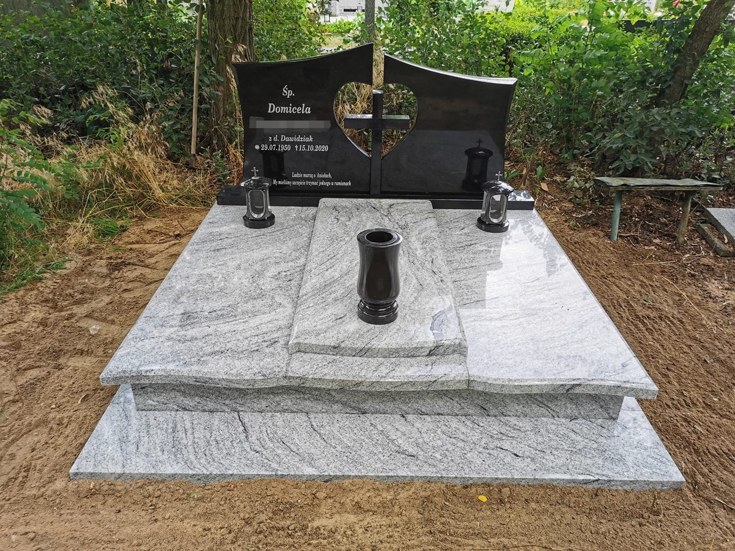 Podwójny, czarny pomnik z wyciętym sercem i jasnoszarą, stopniowaną podstawą, otoczony zielenią i ziemią.