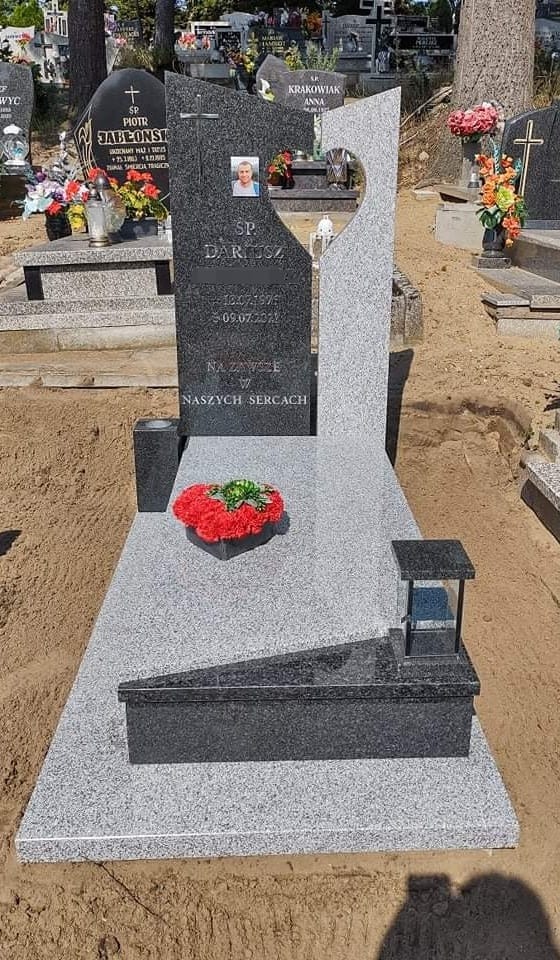 Nowoczesny, dwukolorowy pomnik z ciemnego i jasnego granitu, z wyciętym sercem i zdjęciem zmarłego, udekorowany wieńcem z czerwonych goździków i lampionem.