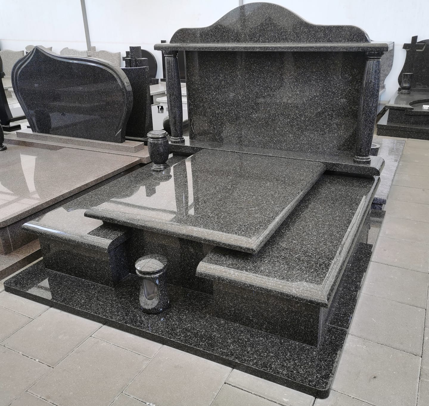 Czarny, granitowy pomnik z kolumnami i dwupoziomową płytą, z widocznymi wazonami i innymi nagrobkami w tle.