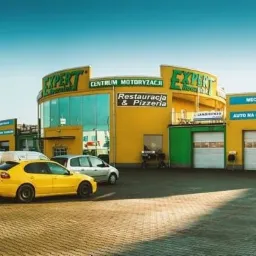 Żółty budynek centrum motoryzacyjnego z restauracją i pizzą, oferującego mechanikę, elektrykę samochodową, serwis ogumienia i myjnię, z zaparkowanymi samochodami na brukowanym placu.
