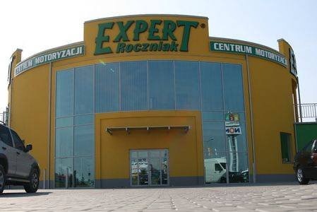 Żółty budynek Centrum Motoryzacji Expert Roczniak z dużymi oknami, widok z zewnątrz na parking z zaparkowanymi samochodami.