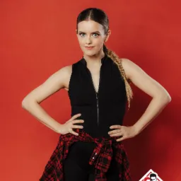 Portret kobiety w czarnym stroju sportowym z rozpiętym suwakiem, z czerwoną koszulą w kratę zawiązaną w pasie, stojącej na tle czerwonej ściany, z logo w prawym dolnym rogu.