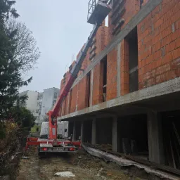 Budowa domu z czerwonej cegły: robotnik na podnośniku układa drewniane elementy. Widoczny fragment elewacji i podnośnik na tle budynku mieszkalnego.