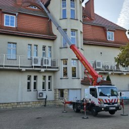 MKM service sp .Z.O.O - Praca na wysokościach przy budynku z użyciem podnośnika koszowego; widoczny pracownik na dachu. Czerwony żuraw na tle jasnej elewacji i okien.