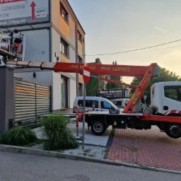 Pracownicy na podnośniku koszowym montują reklamę zewnętrzną na budynku. Widoczny fragment reklamy, budynek, podnośnik i zaparkowany samochód. Dzień, ujęcie z dołu.