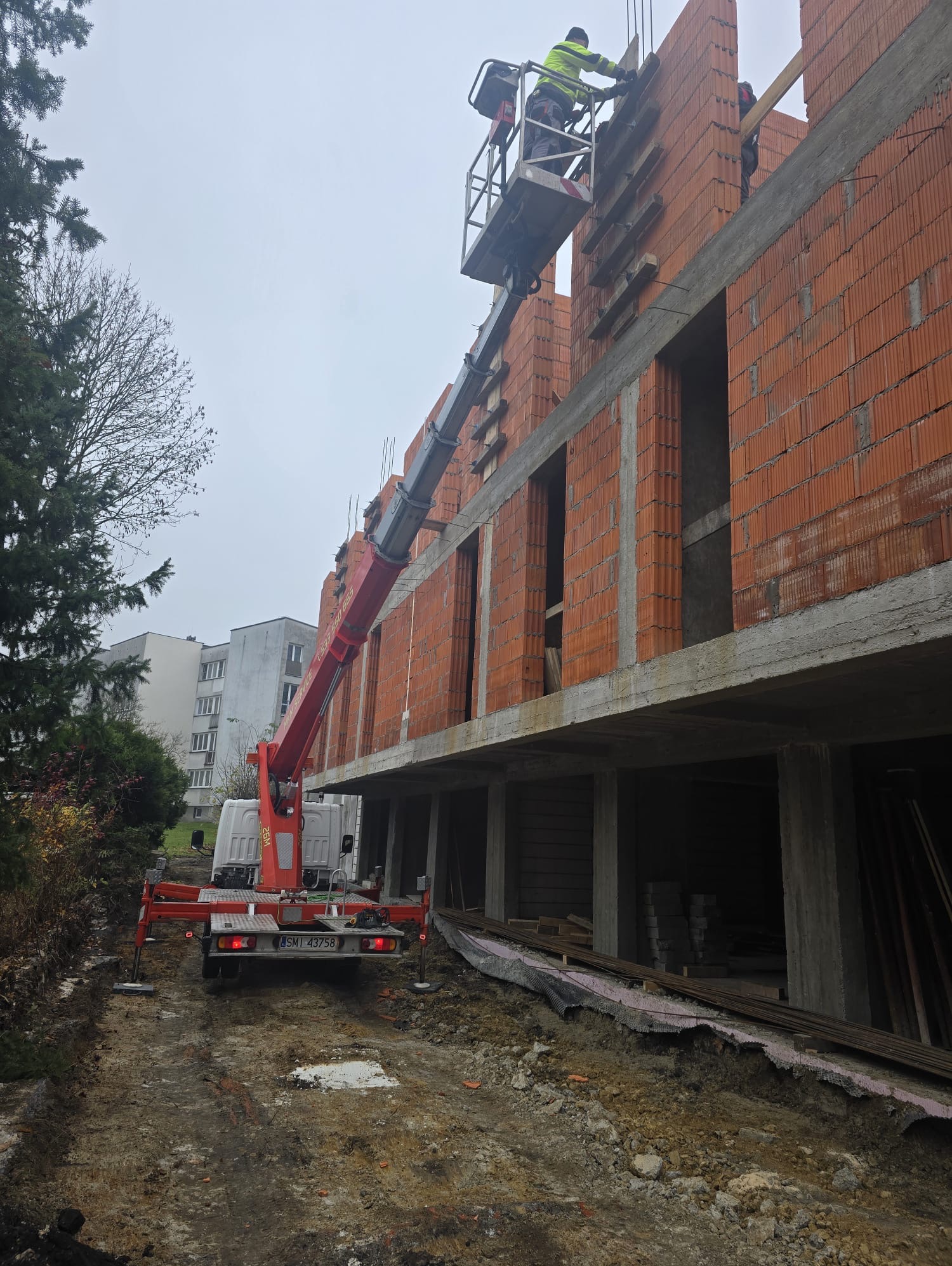 Budowa domu z czerwonej cegły: robotnik na podnośniku układa drewniane elementy. Widoczny fragment elewacji i podnośnik na tle budynku mieszkalnego.