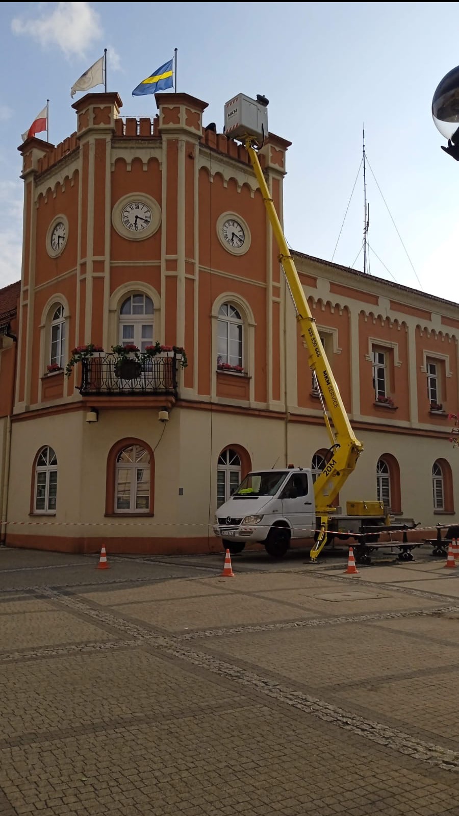 Żółty podnośnik koszowy na tle budynku z zegarami i flagami. Biały bus zaparkowany obok. Pomarańczowe pachołki na brukowanej nawierzchni.