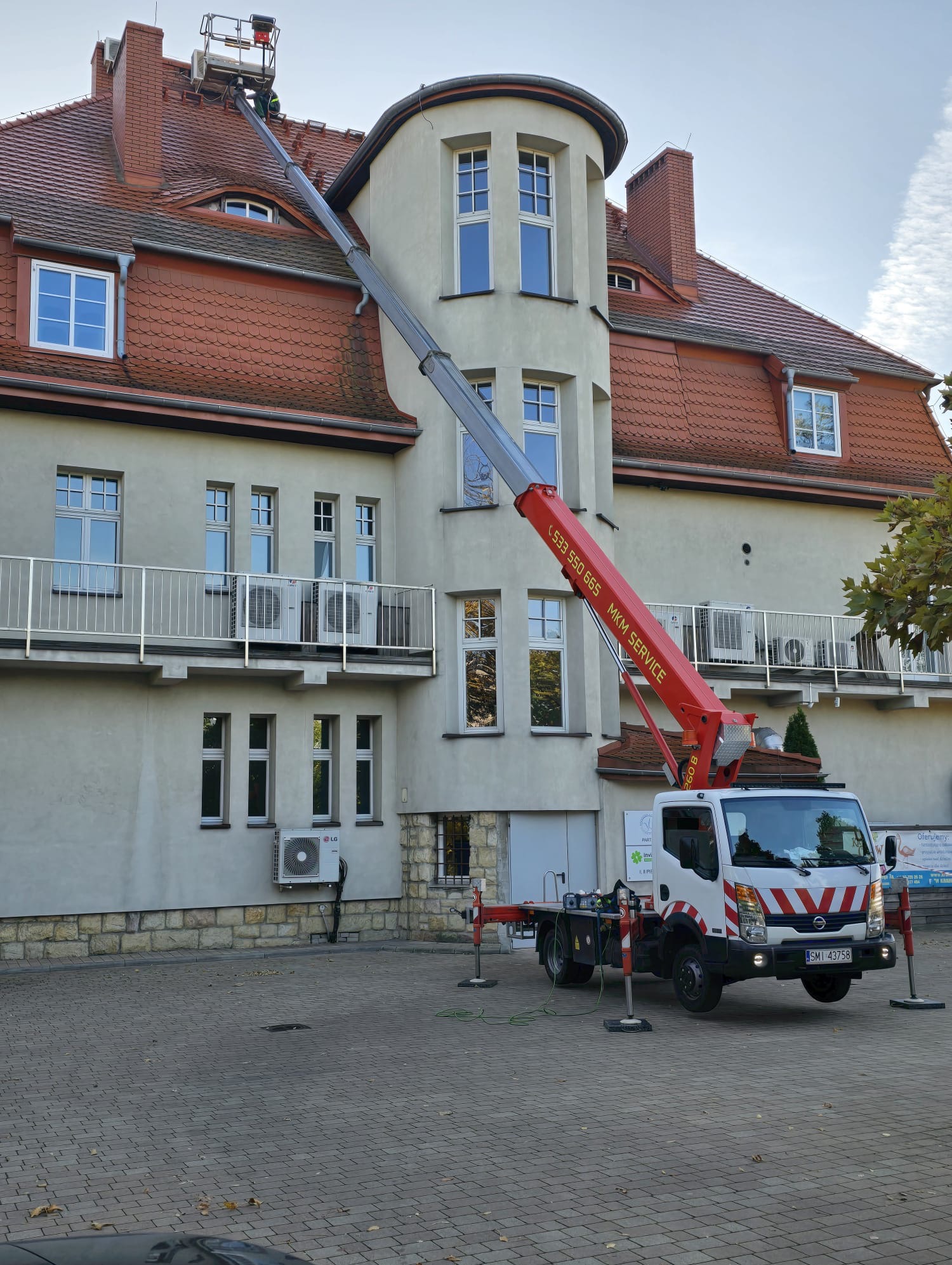 Praca na wysokościach przy budynku z użyciem podnośnika koszowego; widoczny pracownik na dachu. Czerwony żuraw na tle jasnej elewacji i okien.