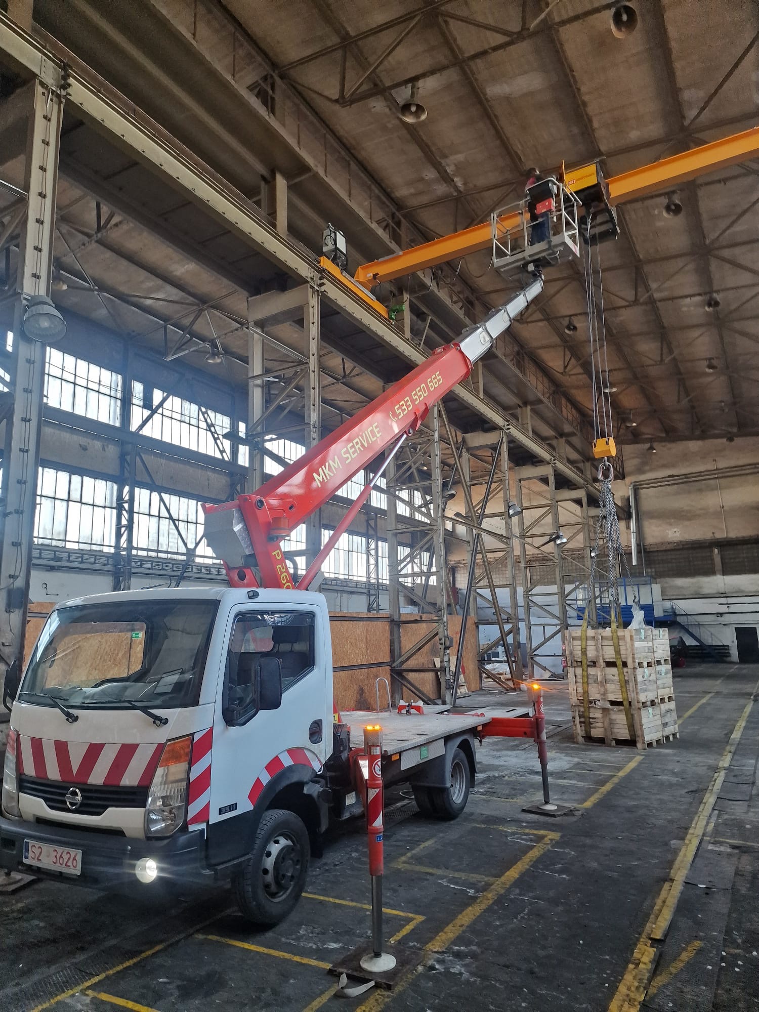 Wnętrze hali przemysłowej: mobilny dźwig z podnośnikiem transportuje paletę. Widoczna konstrukcja hali i okna. Na dźwigu pracownik w uprzęży.