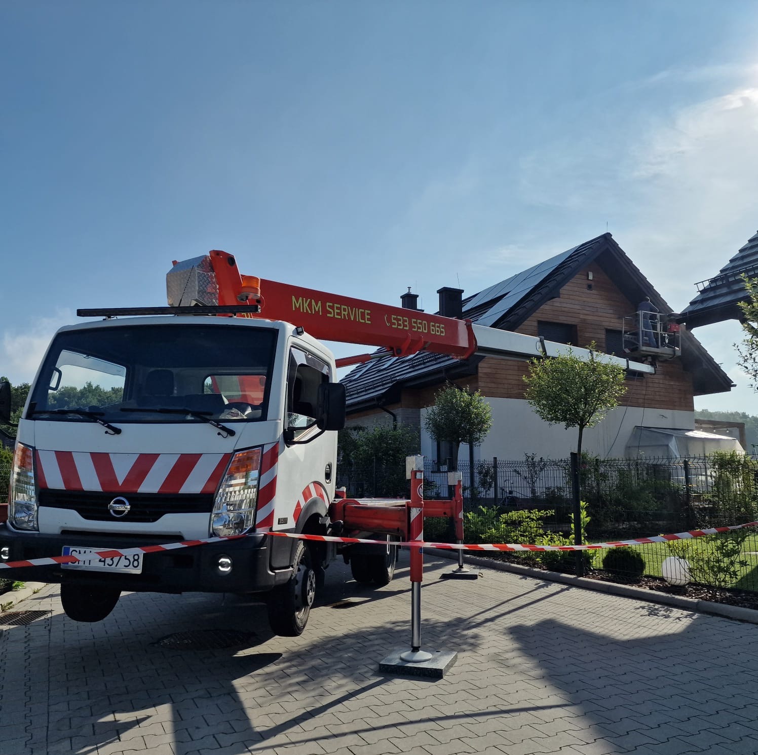 Praca na wysokościach: Podnośnik koszowy MKM Service na tle domu z panelami słonecznymi. Na platformie roboczej pracownik wykonuje zadania. Oznakowany teren.