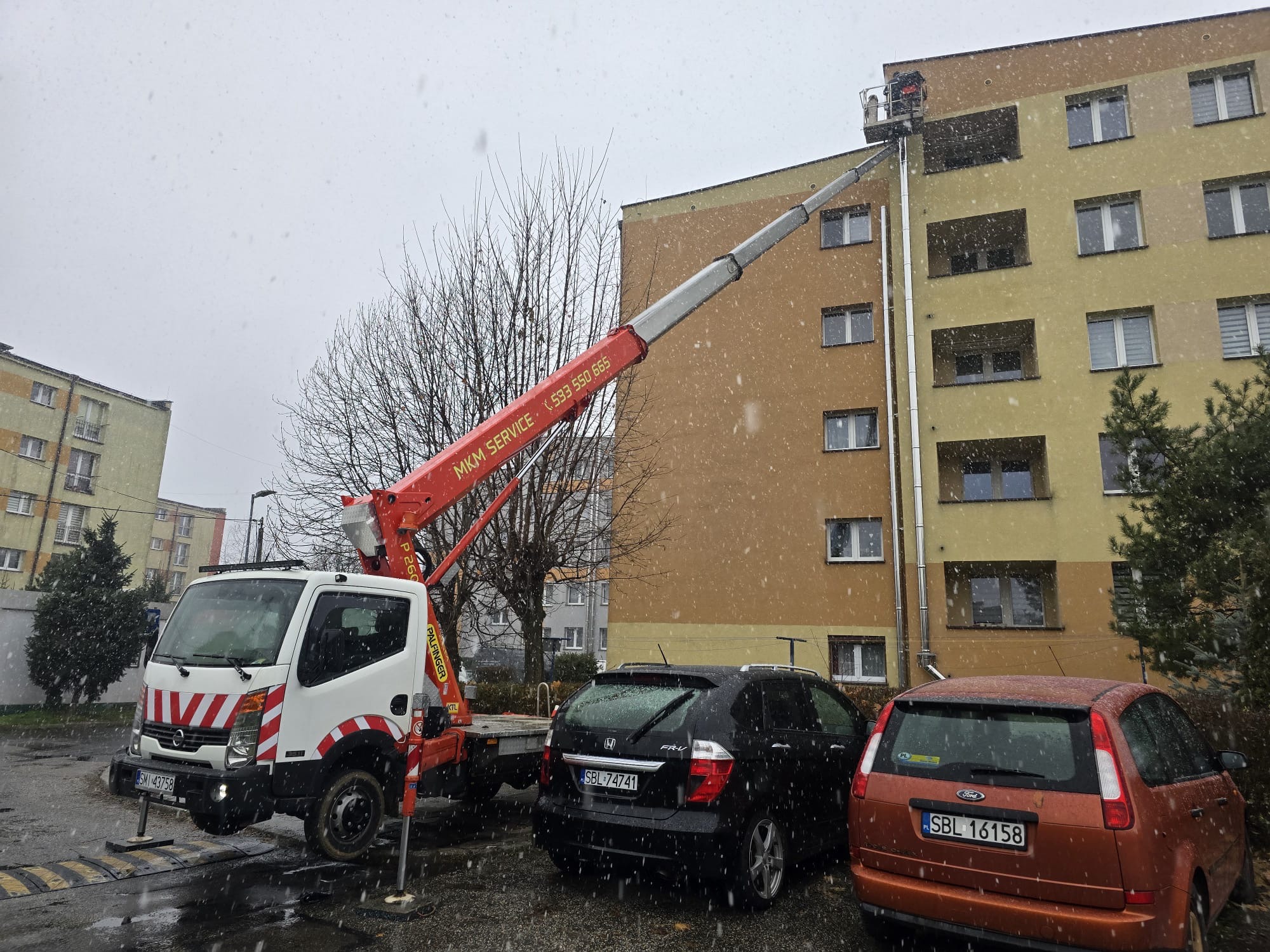 Praca na wysokości: Podnośnik koszowy na tle bloku podczas opadów śniegu. Widoczny pracownik w koszu. Na pierwszym planie zaparkowane auta.