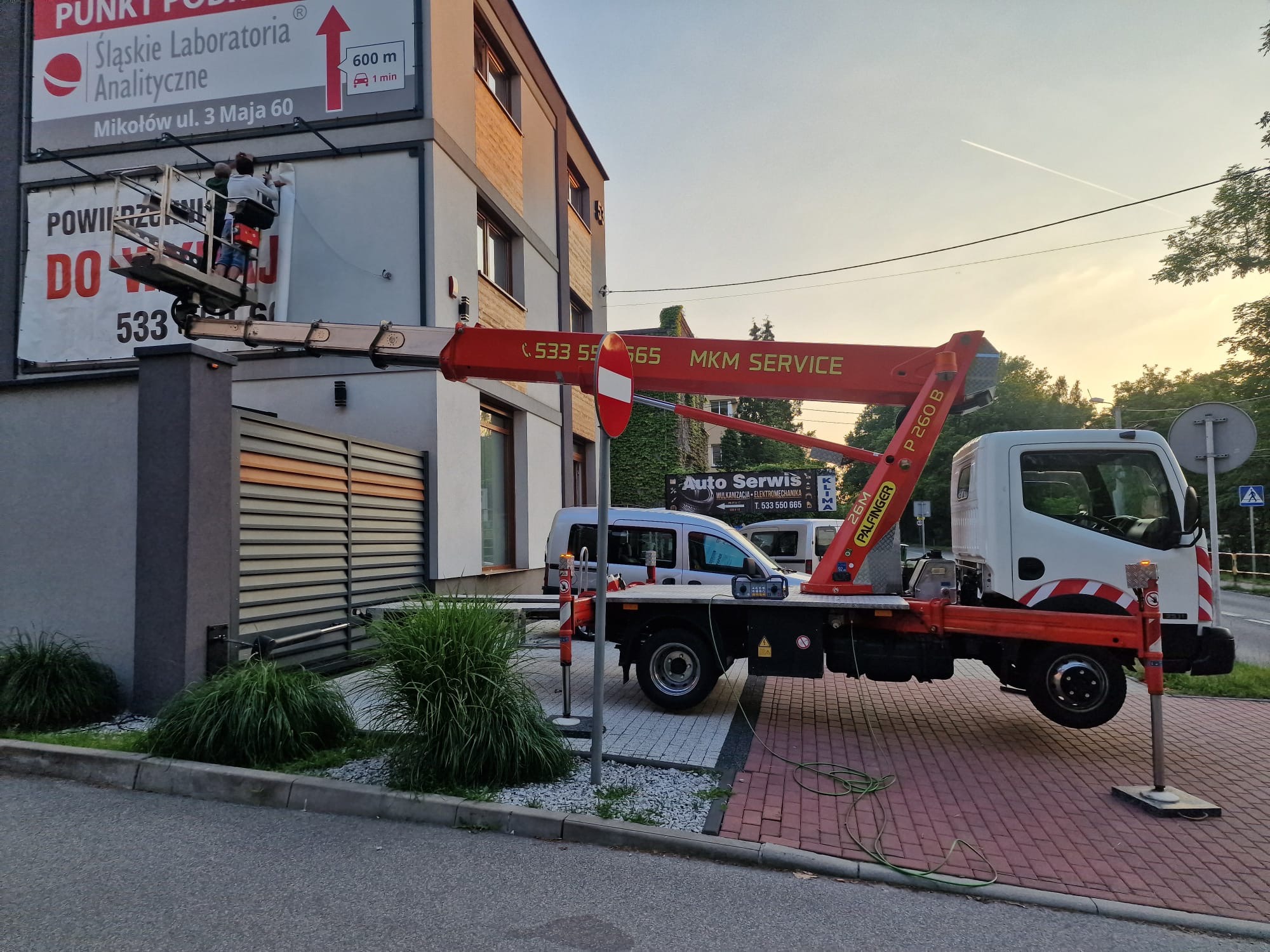 Pracownicy na podnośniku koszowym montują reklamę zewnętrzną na budynku. Widoczny fragment reklamy, budynek, podnośnik i zaparkowany samochód. Dzień, ujęcie z dołu.