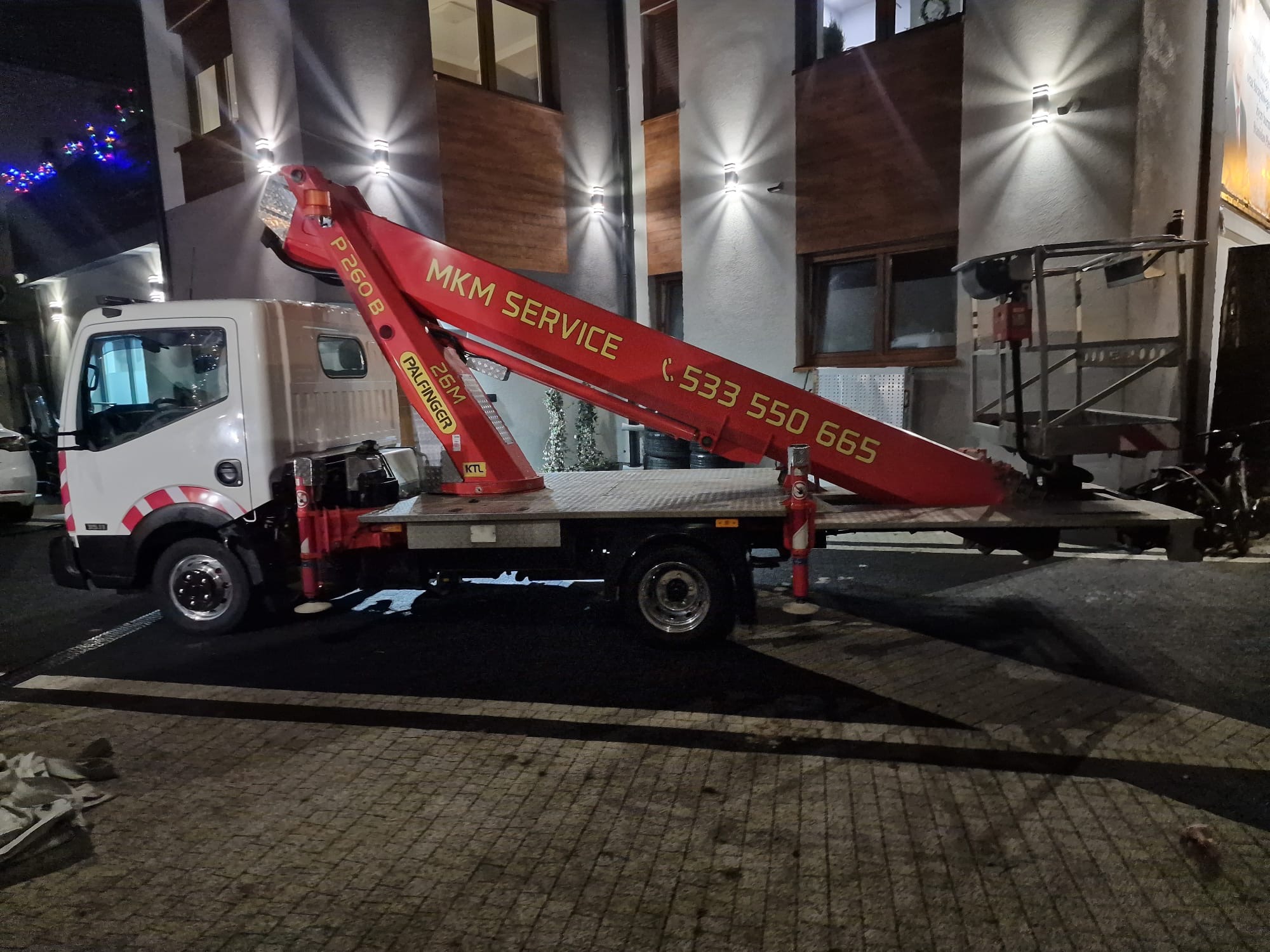 Podnośnik koszowy MKM Service z numerem telefonu na tle oświetlonej elewacji budynku. Widoczne elementy dekoracji świątecznej.