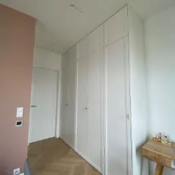 Biała szafa wnękowa z złotymi uchwytami, dopasowana do skosu sufitu, z parkietem w jodełkę i toaletką w rogu pokoju, ściana w kolorze pudrowego różu.