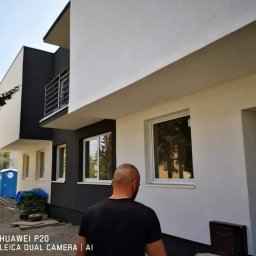 Usługi remontowo budowlane - Dwukondygnacyjny budynek z biało-szarą elewacją po remoncie, widoczny fragment ulicy z przenośną toaletą i stertą piasku, w kadrze osoba w czarnej koszulce.