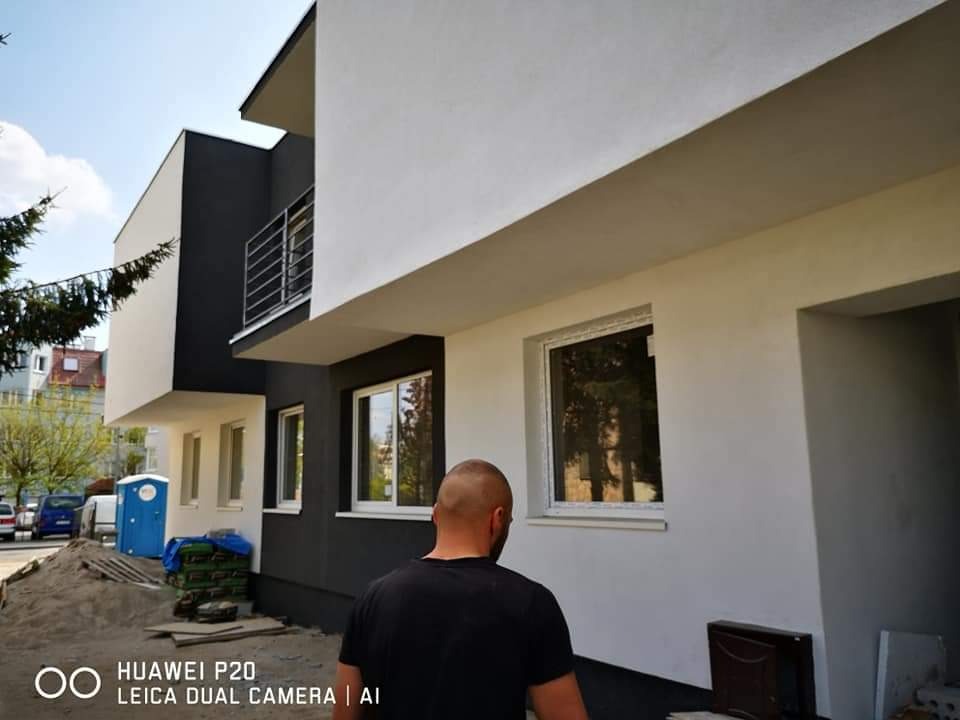 Dwukondygnacyjny budynek z biało-szarą elewacją po remoncie, widoczny fragment ulicy z przenośną toaletą i stertą piasku, w kadrze osoba w czarnej koszulce.