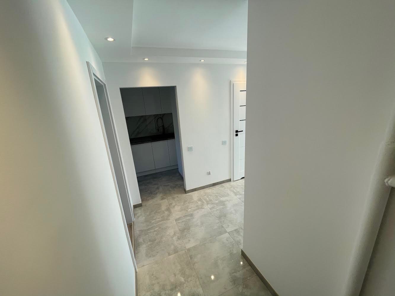 Wnętrze mieszkania po remoncie: białe ściany, szara podłoga z płytek, widoczny fragment kuchni z białymi szafkami i czarnym blatem, białe drzwi z czarną klamką i akcentami.